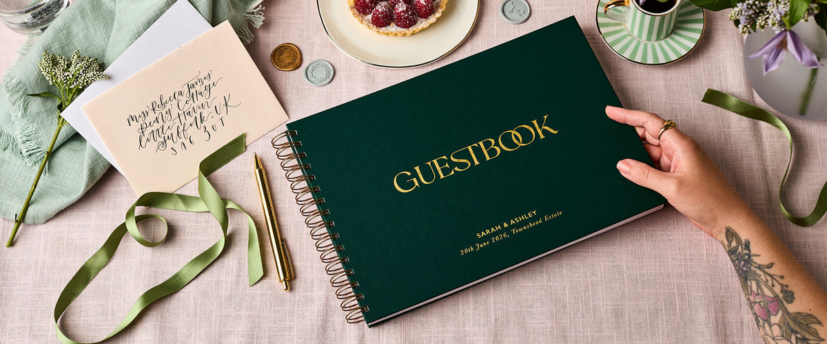 Martha Brook Personalised Guest Book in green on pink fabric next to envelopes and ribbon
