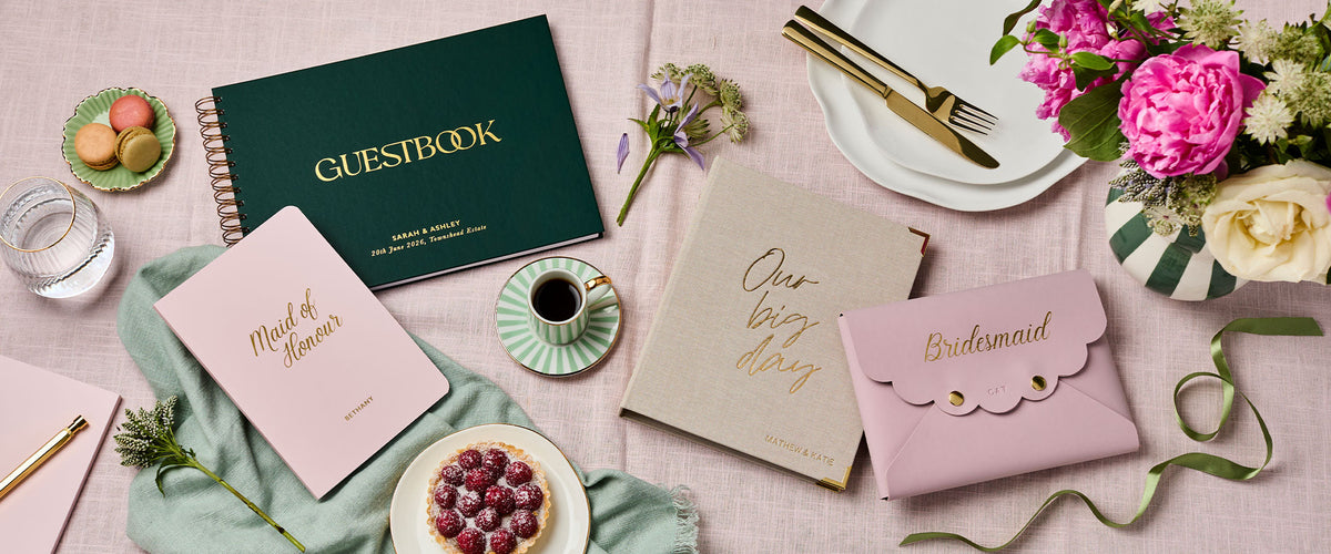 Martha Brook Personalised Wedding Planner, Personalised Guest Book, Personalised Maid of Honour Softback Notebook, Personalised Bridesmaid Pouch on a pink table with flowers and ribbons