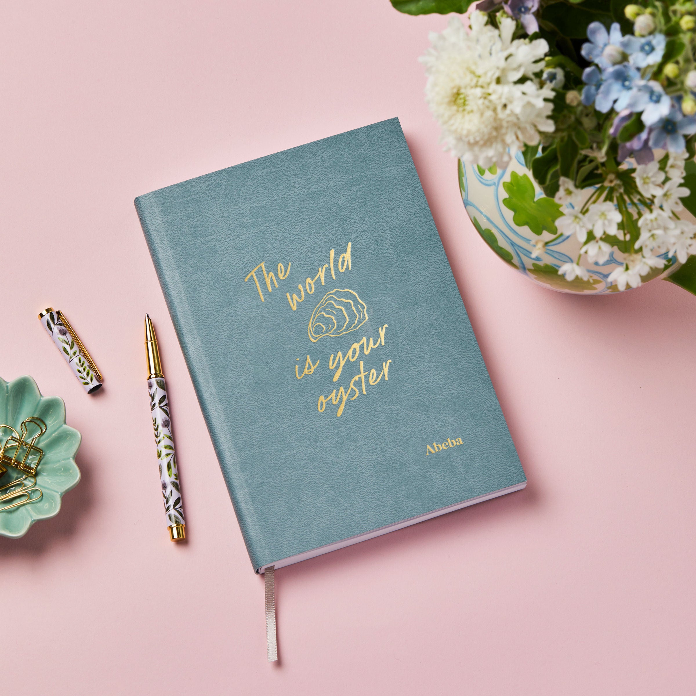 Martha brook personalised stationery personalised leaving gift vegan leather notebook teal notebook with gold foil reading 'the world is your oyster' on a pink background