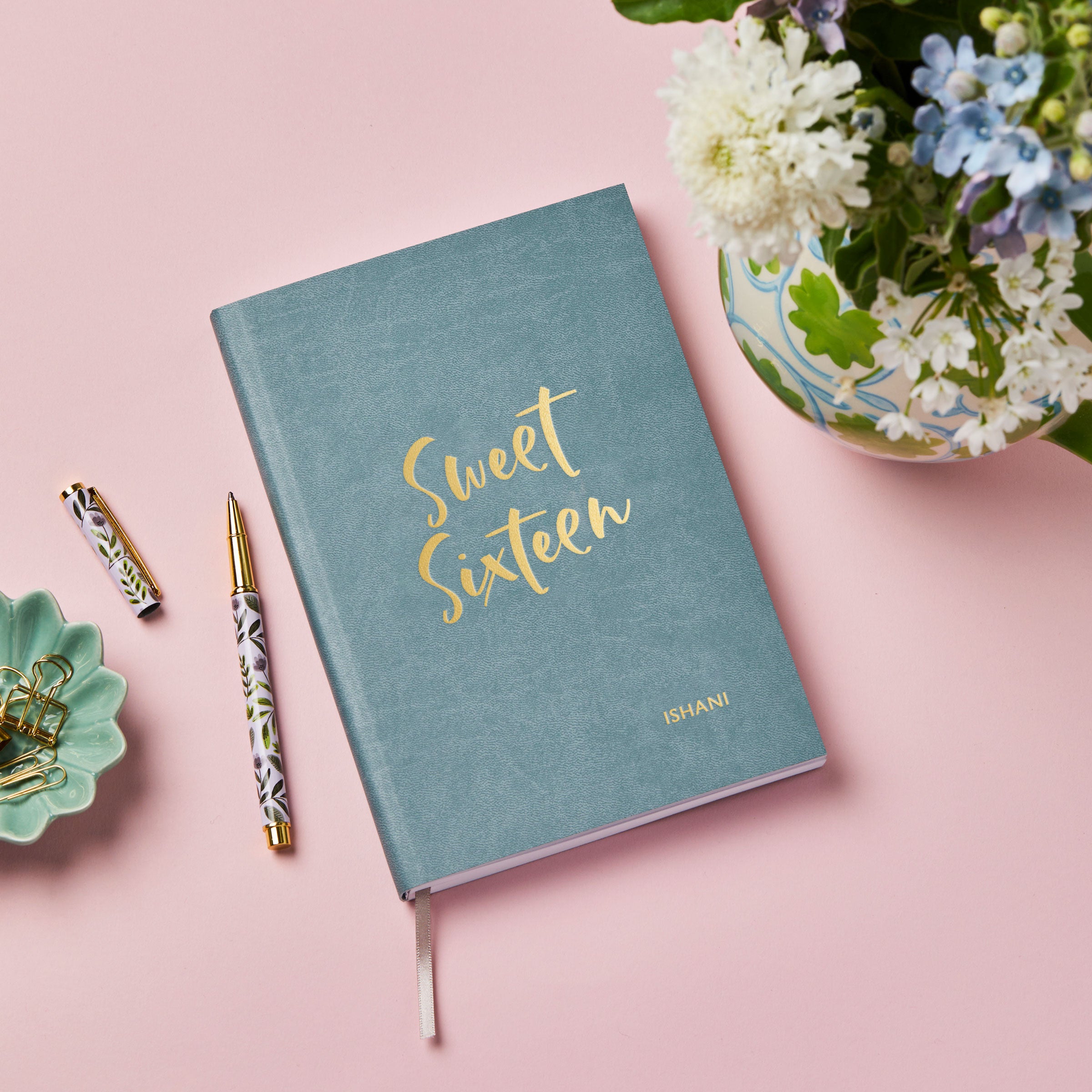martha brook personalised stationery personalised 16th birthday gift notebook main image. A teal blue vegan leather notebook on a pink desk/backdrop. The cover of the notebook reads 'sweet sixteen' in gold foil