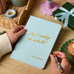 Martha Brook personalised stationery personalised 18th birthday gift softback notebook main image. A pale blue notebook on a wooden desk. The notebook reads 'Am I really an adult? in gold foiling