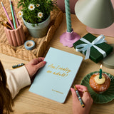Martha Brook personalised stationery personalised 18th birthday gift softback notebook main image. A pale blue notebook on a wooden desk. The notebook reads 'Am I really an adult? in gold foiling. On the desk around there is a present wrapped in green paper with a blue ribbon, and a muffin with a candle in it