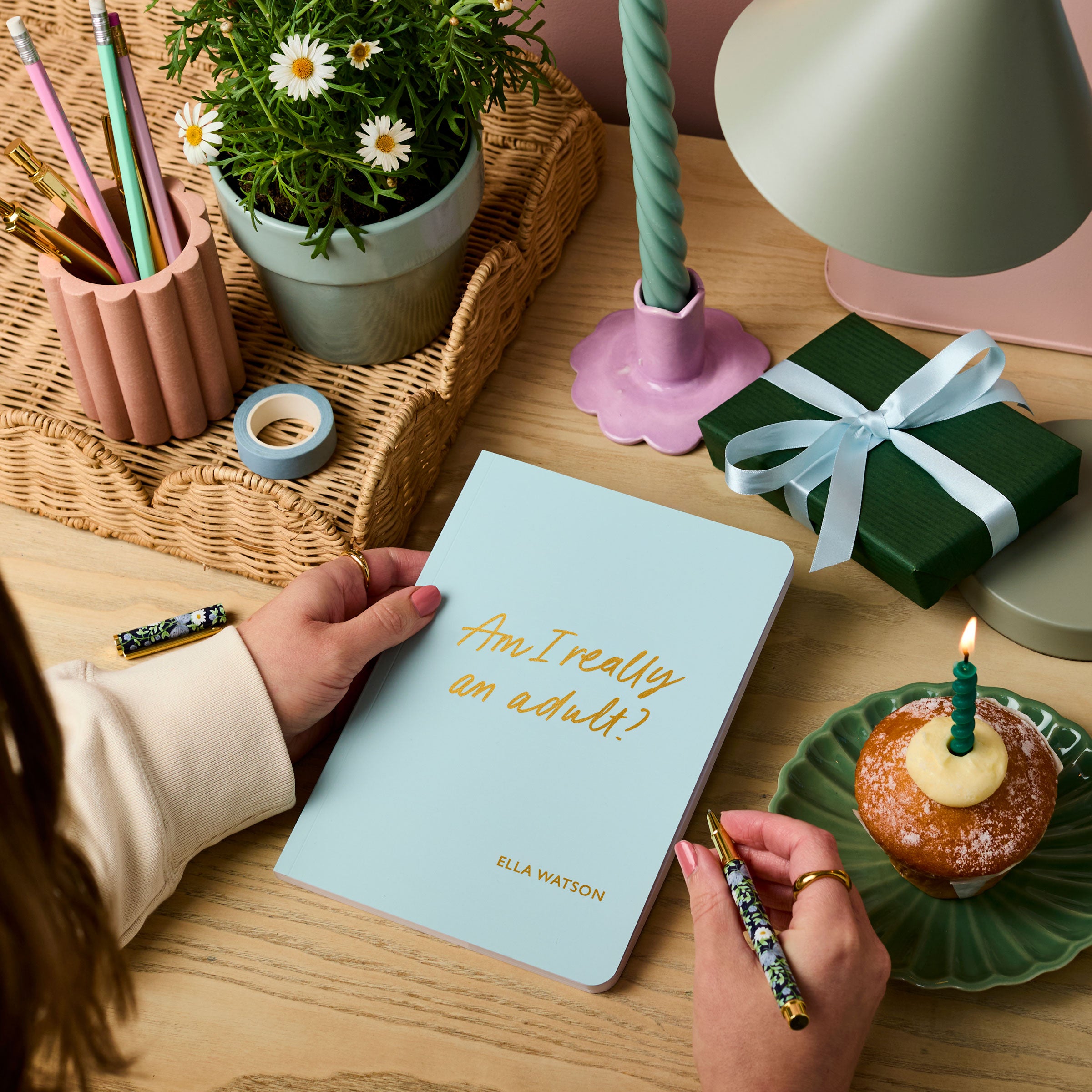 Martha Brook personalised stationery personalised 18th birthday gift softback notebook main image. A pale blue notebook on a wooden desk. The notebook reads 'Am I really an adult? in gold foiling. On the desk around there is a present wrapped in green paper with a blue ribbon, and a muffin with a candle in it