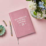 martha brook personalised stationery personalised 21st birthday gift softback notebook. A pink vegan leather notebook on a pink background reads 'it's supposed to be fun turning 21' in silver foiling on the cover. To it's left is a small green trinket tray filled with gold bulldog clips, and in the top right is a vase of spring flowers