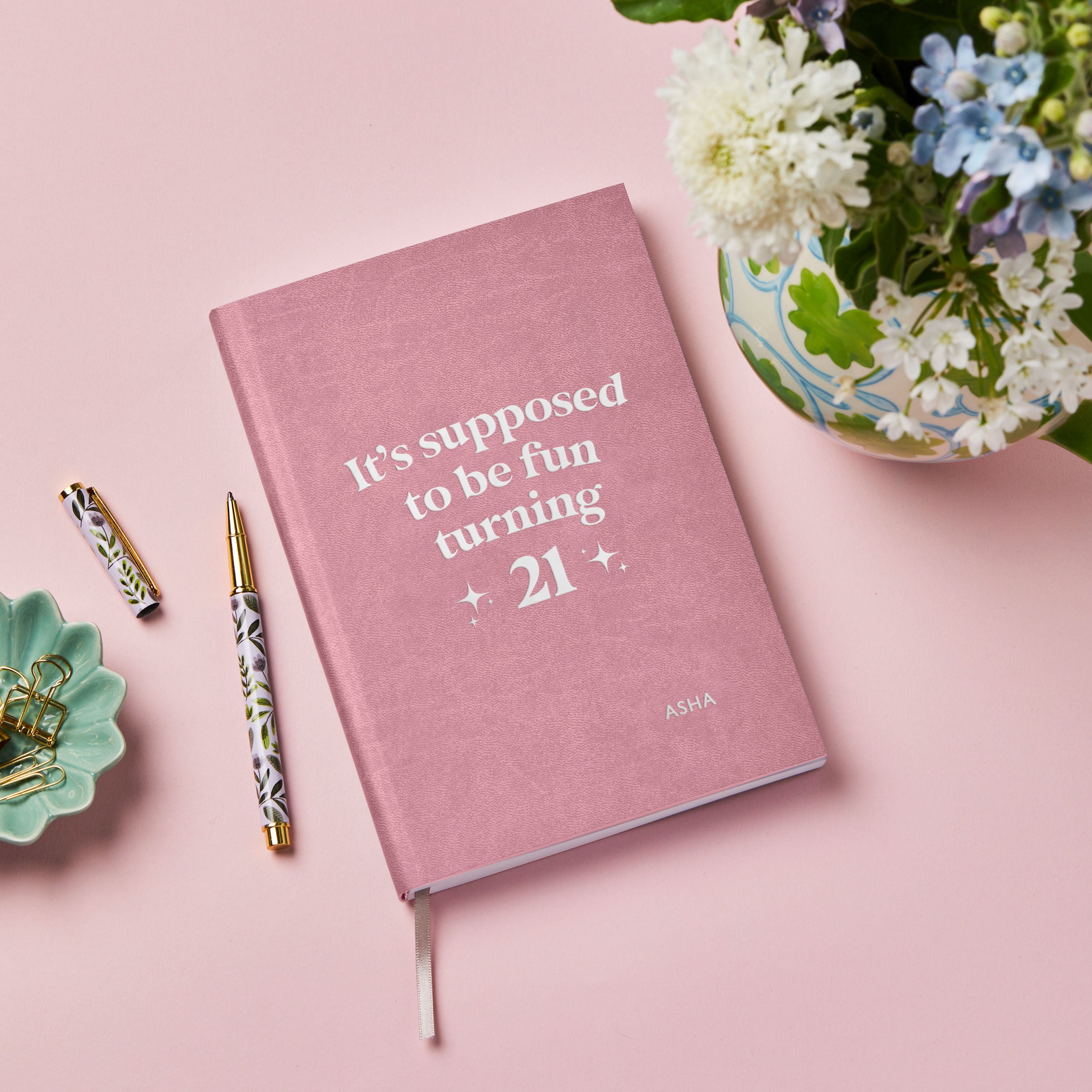 martha brook personalised stationery personalised 21st birthday gift softback notebook. A pink vegan leather notebook on a pink background reads 'it's supposed to be fun turning 21' in silver foiling on the cover. To it's left is a small green trinket tray filled with gold bulldog clips, and in the top right is a vase of spring flowers