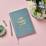 martha brook personalised stationery personalised 40th birthday gift notebook light blue notebook on a wooden desk. The notebook reads 'life beings at 40. Up until then you're just doing research' in gold foil on the cover