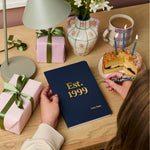 Martha Brook personalised stationery personalised any year birthday softback notebook navy blue notebook on desk with 'est. 1999' embossed on the cover in gold foil with pink birthday presents wrapped in green ribbon on the desk. A victoria sponge birthday cake with candles is on the desk next to the notebook