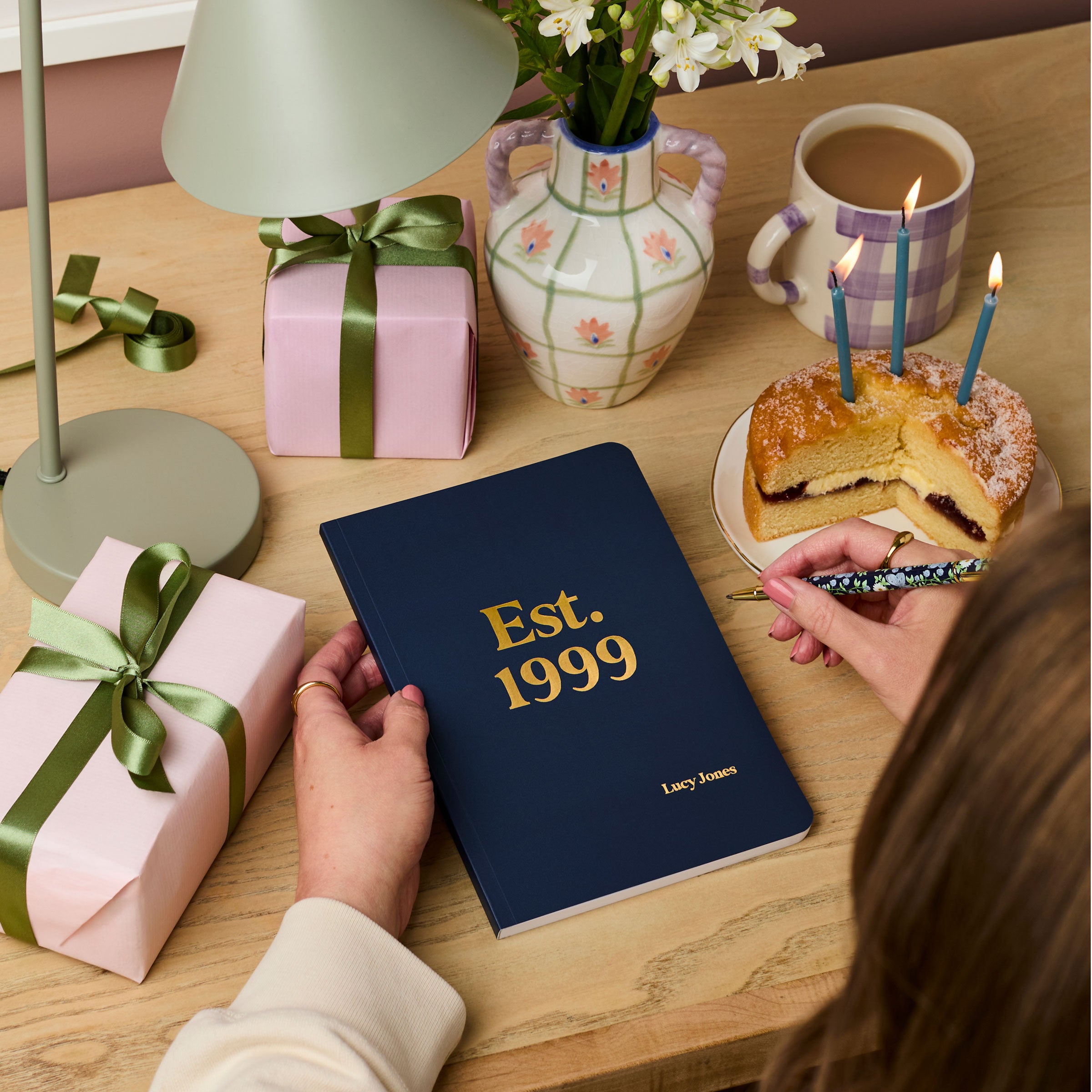 Martha Brook personalised stationery personalised any year birthday softback notebook navy blue notebook on desk with 'est. 1999' embossed on the cover in gold foil with pink birthday presents wrapped in green ribbon on the desk. A victoria sponge birthday cake with candles is on the desk next to the notebook