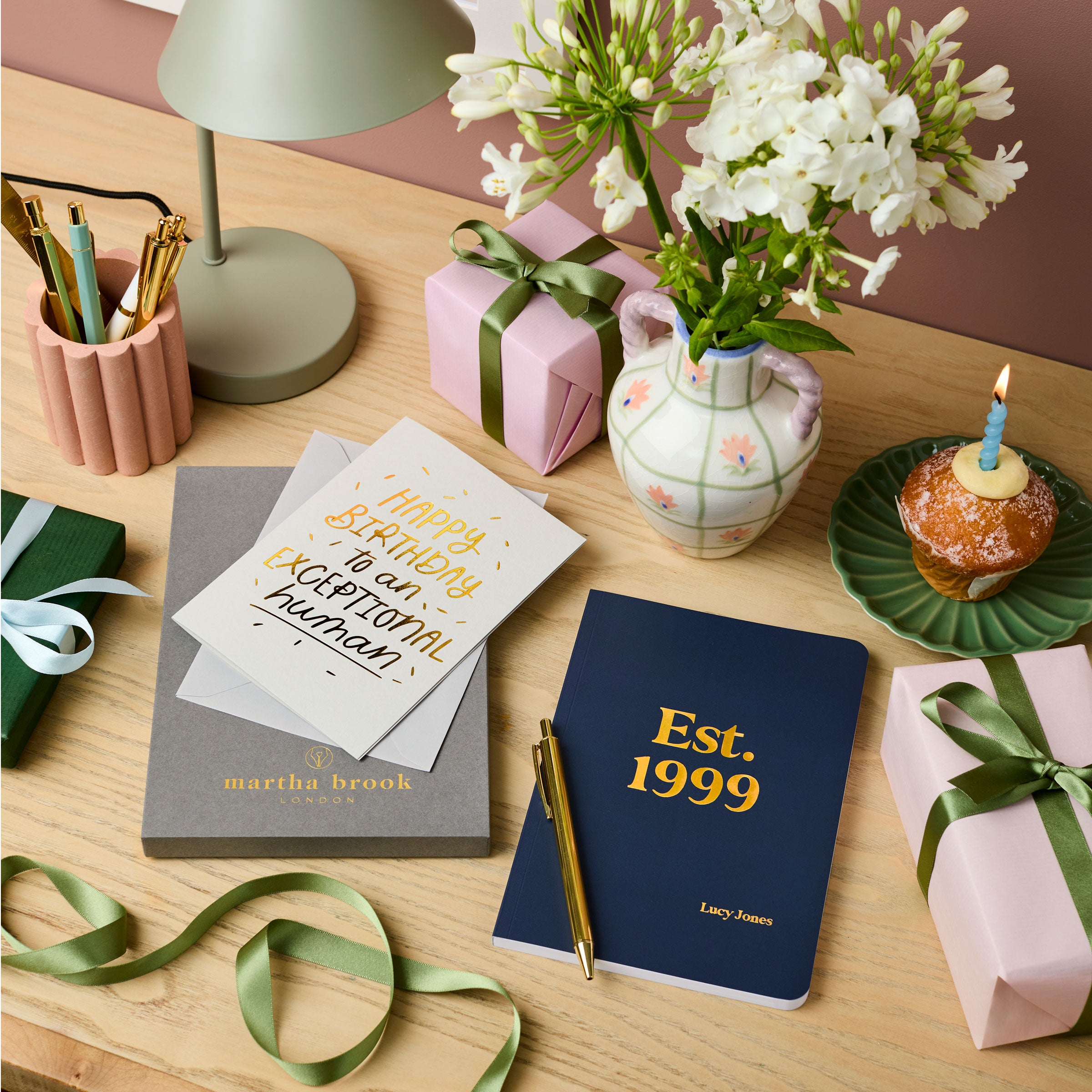 martha brook personalised stationery personalised birthday gift set. A grey gift box, a birthday card reading 'Happy Birthday to an exceptional human', a navy blue notebook reading 'est. 1999' in gold foil on the cover, and a gold pen are all laid out on a desk.