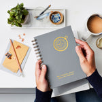 martha brook personalised stationery personalised compass luxe hardback notebook in slat grey with gold foil on a coffee table with a mug of tea and marble tray