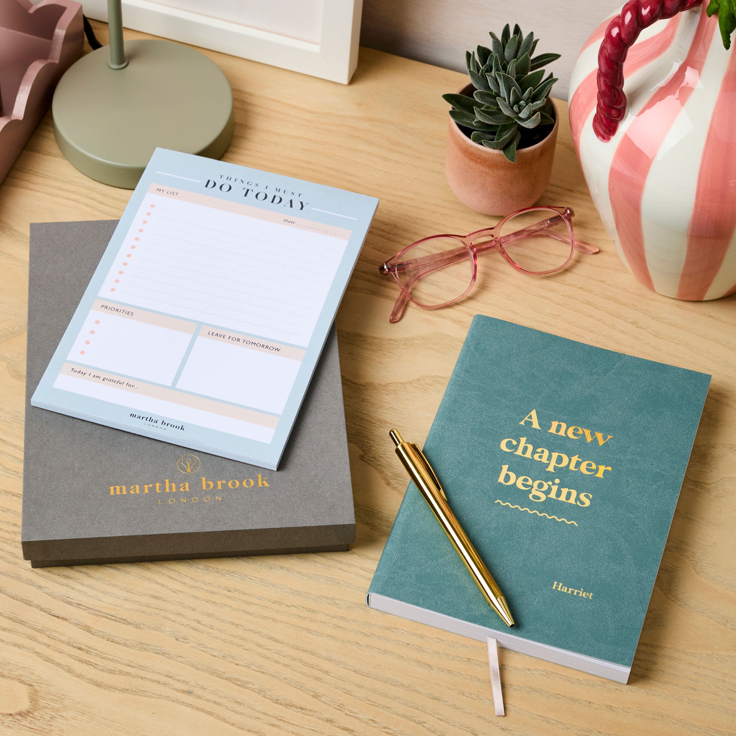 martha brook personalised stationery personalised new job gift set. A grey gift box, a desk pad, a teal blue notebook reading 'a new chapter begins' in gold foil on the cover, and a gold pen are all laid out on a desk.
