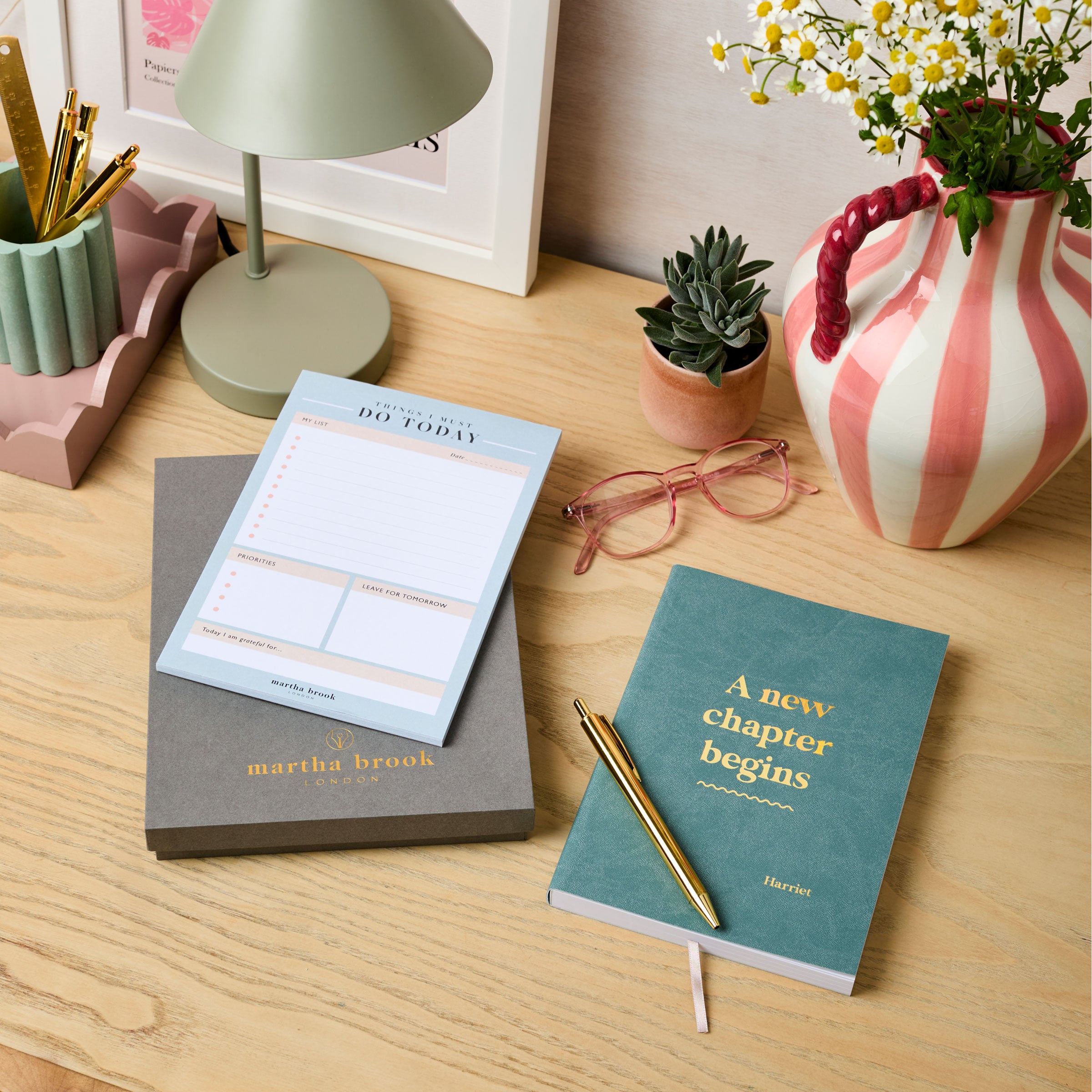 martha brook personalised stationery personalised new job gift set. A grey gift box, a desk pad, a teal blue notebook reading 'a new chapter begins' in gold foil on the cover, and a gold pen are all laid out on a desk.