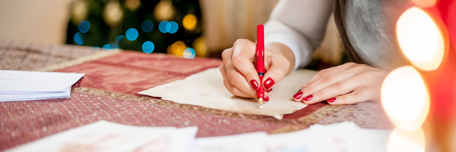 martha brook blog post what to write in a christmas card 30 christmas card messages person writing in a christmas card on a table next to a christmas tree