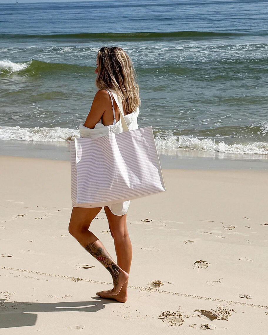 martha brook personalised stationery blog 15 things to take on holiday that you’ll wish you’d thought of sooner sunnylife beach tote bag. A woman walks down the beach holding a large pink and white striped tote bag