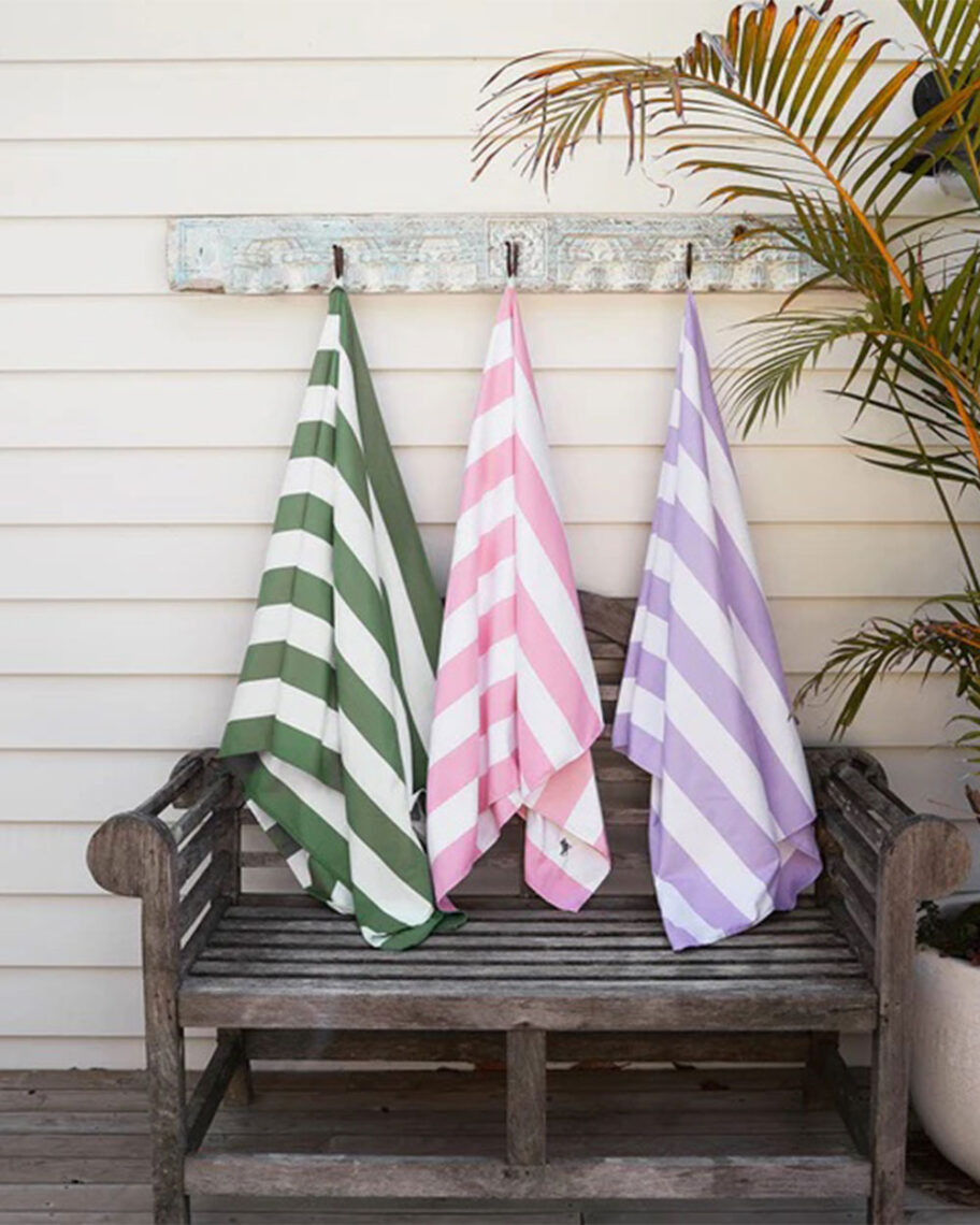 martha brook personalised stationery blog 15 things to take on holiday that you’ll wish you’d thought of sooner dock and bay towel. Three striped towels hang up on hooks outside. One towel is green, one is pink and one is purple. There is a wooden bench underneath them