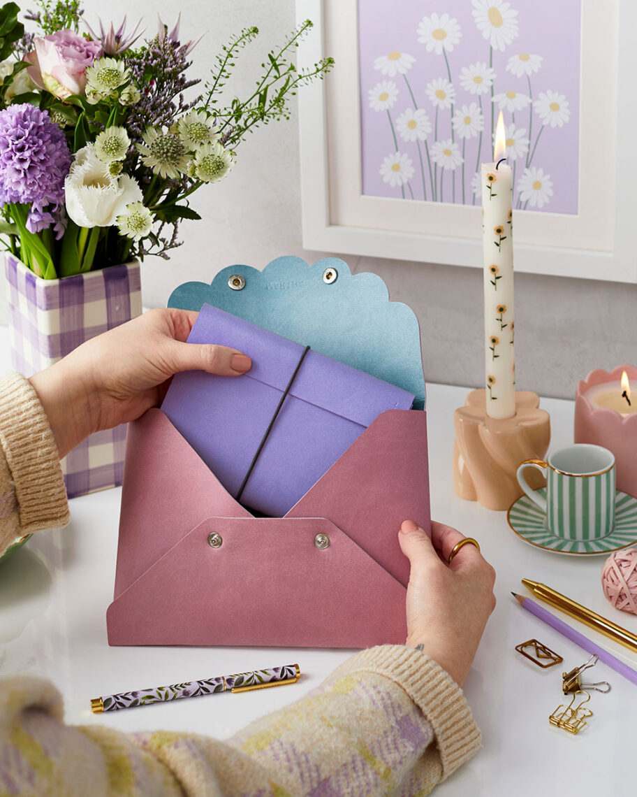 martha brook personalised stationery blog 15 things to take on holiday that you’ll wish you’d thought of sooner a document pouch. A person sat at a desk puts a lilac notebook into a dusky pink pouch with a blue lining