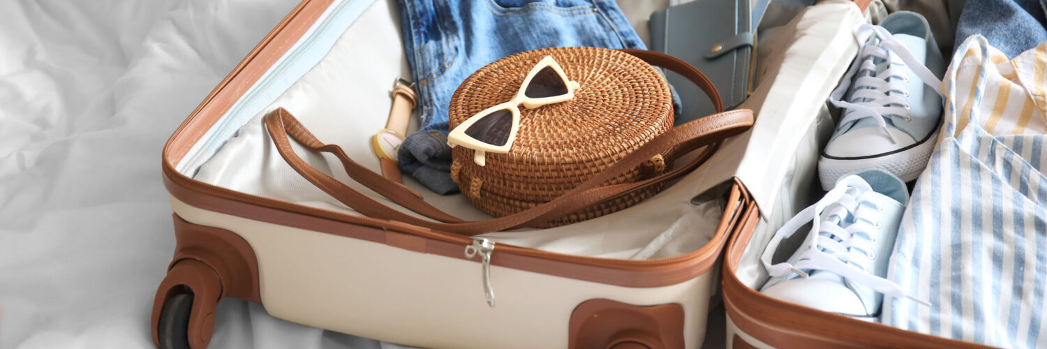 martha brook personalised stationery blog 15 things to take on holiday that you’ll wish you’d thought of sooner open suitcase on a bed. The suitcase is cream with a brown border and wheels. Inside the suitcase are lots of light and neutral coloured cloths, and on top sits a brown woven bag and a pair of sunglasses