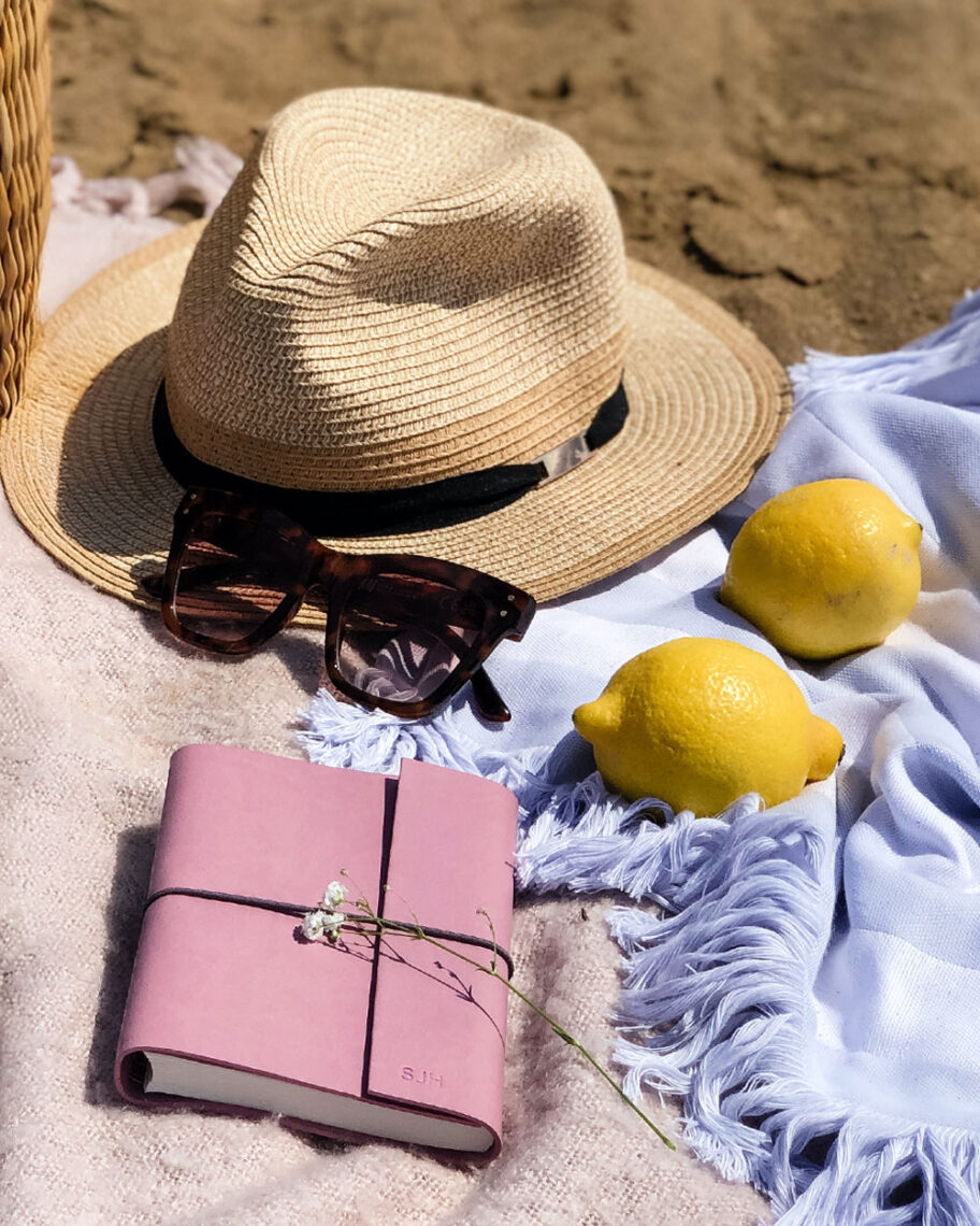 martha brook personalised stationery blog 15 things to take on holiday that you’ll wish you’d thought of sooner personalised vegan leather notebook in dusky rose. A pink notebook, two lemons, a hat and some sunglasses are set out on a blanket on some sand
