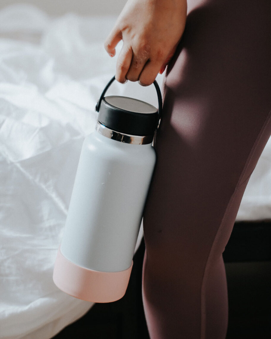 martha brook personalised stationery blog 15 things to take on holiday that you’ll wish you’d thought of sooner empty water bottle. A person wearing brown gym leggings holds a large metal reusable water bottle down by their side. The bottle is pale blue with a stripe of pink at the bottom