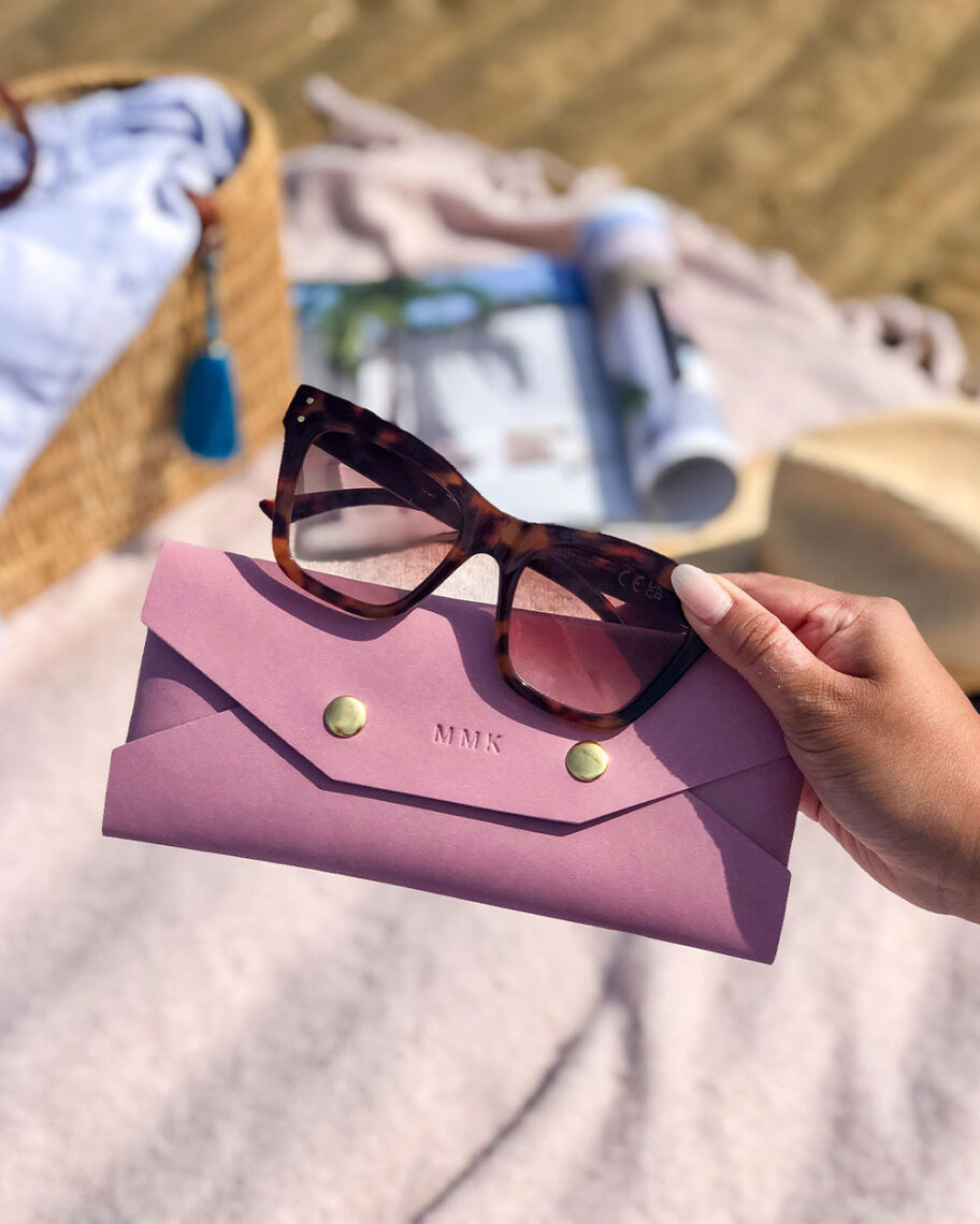 martha brook personalised stationery blog 15 things to take on holiday that you’ll wish you’d thought of sooner personalised vegan leather sunglasses case. A person holds a pink vegan leather sunglasses case and a pair of sunglasses in one hand. In the background a blanket is laid out on the sand