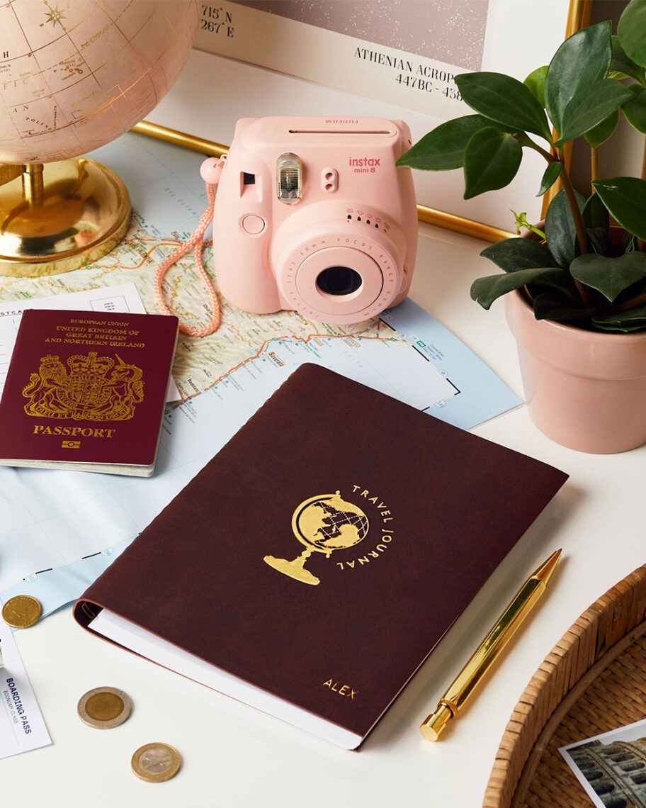 martha brook personalised stationery blog 15 things to take on holiday that you’ll wish you’d thought of sooner personalised travel journal. A burgundy book with a picture of a globe and the words 'travel journal' embossed in gold on the front, sits on a desk surrounded by travel related memorabilia such as a globe, passport, camera and map.
