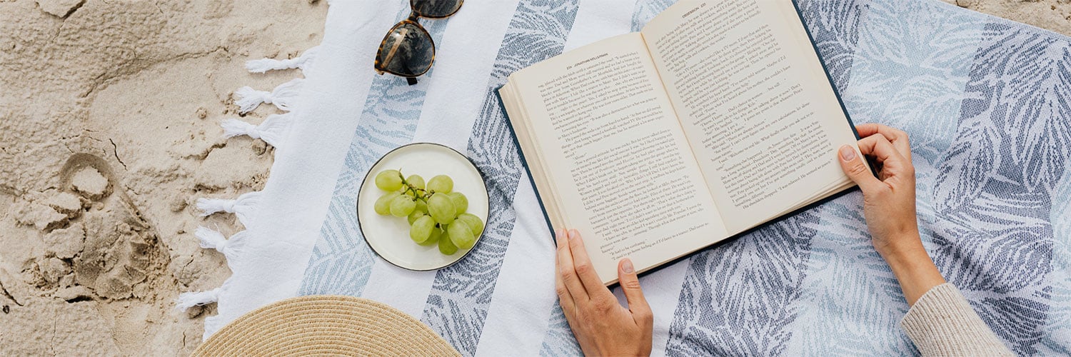 Martha Brook personalised stationery blog 20 best summer reads from old classics to new releases. A person lying on a picnic blanket on a beach with a book open. Next to them they have their sunglasses and a bunch of grapes on a plate