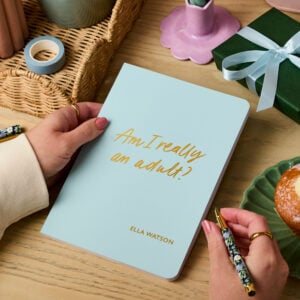 Martha Brook personalised stationery personalised 18th birthday gift softback notebook main image. A pale blue notebook on a wooden desk. The notebook reads 'Am I really an adult? in gold foiling