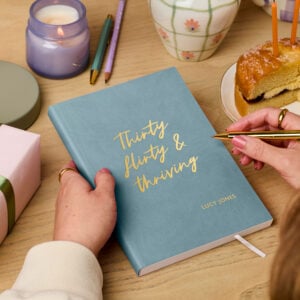 martha brook personalised stationery personalised 30th birthday gift notebook. A teal vegan leather notebook on a wooden desk reads 'thirty, flirty and thriving' on the cover in gold foil