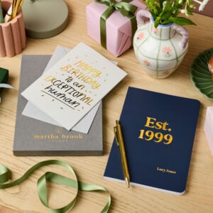 martha brook personalised stationery personalised birthday gift set. A grey gift box, a birthday card reading 'Happy Birthday to an exceptional human', a navy blue notebook reading 'est. 1999' in gold foil on the cover, and a gold pen are all laid out on a desk.