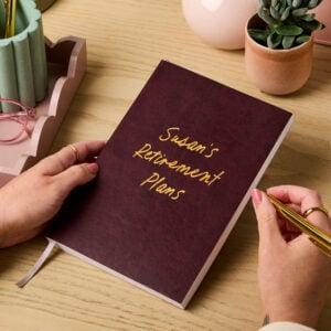 martha brook personalised stationery personalised retirement gift notebook. A burgundy vegan leather notebook on a wooden desk reads 'susan's retirement plans' on the cover in gold foil