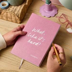 martha brook personalised stationery personalised university gift softback notebook pink vegan leather notebook on a wooden desk. The notebook reads 'what, like it's hard?' on the cover in silver foil