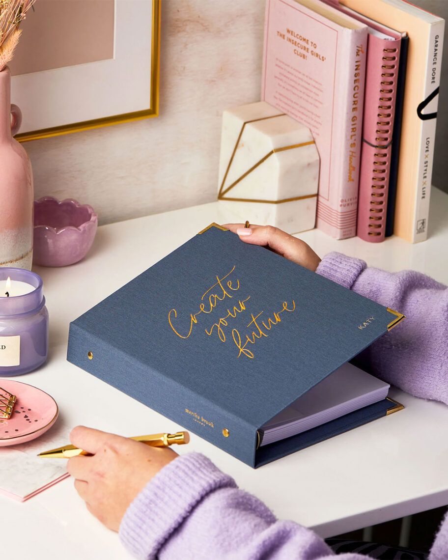 Martha Brook personalised stationery blog 10 dreamy school supplies fror grown-ups that give us that back to school feeling create your future life planner person wearing a purple jumper sat at a desk about to open a blue planner that says 'create your future' in gold embossed script on the front.