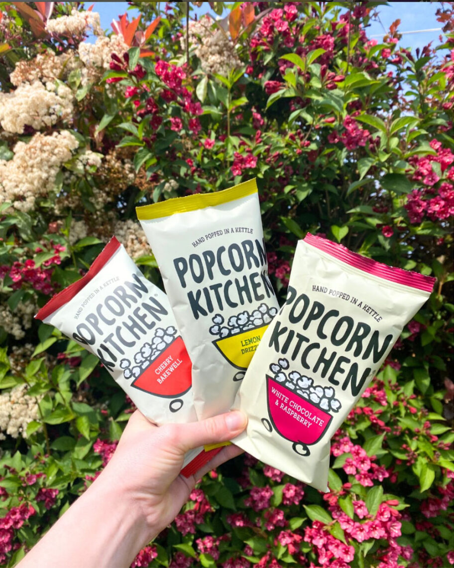 Martha Brook personalised stationery blog 10 dreamy school supplies fror grown-ups that give us that back to school feeling popcorn kitchen mini snack bags. A person in a garden holding up three mini bags of popcorn kitchen popcorn in front of a bush with pink flowers on it