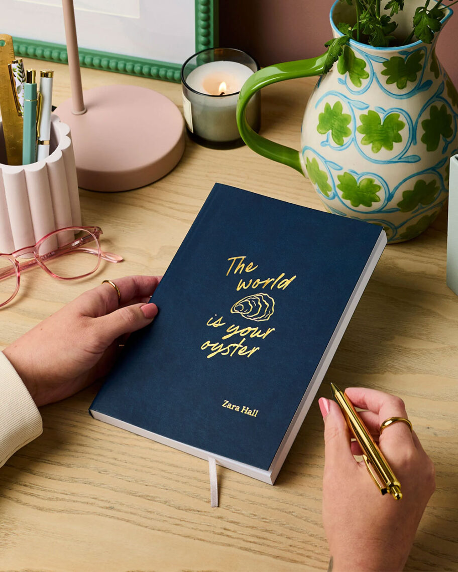 Martha Brook personalised stationery blog 10 dreamy school supplies for grown-ups that give us that back to school feeling the world is your oyster notebook person holding a navy blue notebook on a desk. The notebook says 'the world is your oyster' in gold embossed foil and has a small illustration of an oyster in the middle.