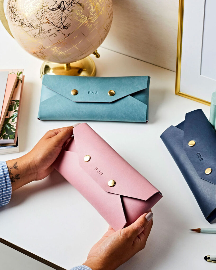 Martha Brook personalised stationery blog 10 dreamy school supplies fror grown-ups that give us that back to school feeling martha brook vegan leather personalised pencil case. Person holding a pink vegan leather pencil case on a desk with two other pencil cases, one light blue and one dark blue. The pencil cases have two gold poppers on the front and are embossed with initials