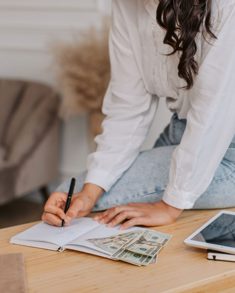 Martha Brook personalised stationery blog Our university essentials and top tips if you’re heading off to study this year girl sat at table writing in a notebook with a pile of money next to her, budgeting