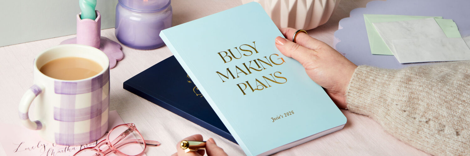 martha brook personalised stationery personalised weekly 2026 diaries. A light blue softback diary on a desk that reads 'busy making plans' in gold foil on the front cover.