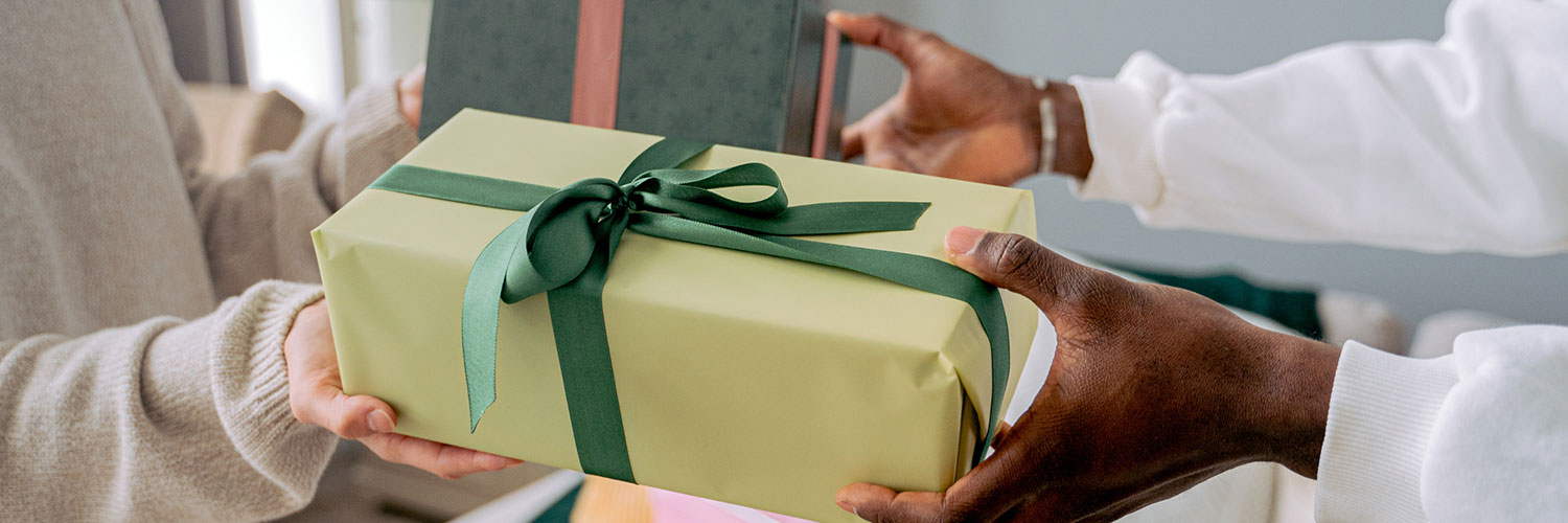 martha brook personalised stationery blog 10 unique birth year personalised birthday gifts header. One person handing a gift to another. The gift is wrapped in light green paper and tied with a dark green ribbon.