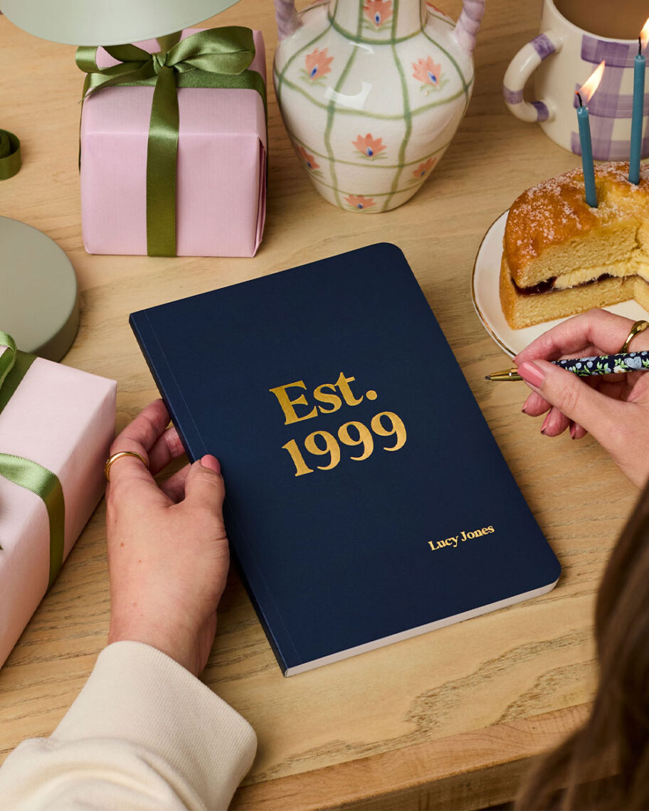 martha brook personalised stationery blog 10 unique birth year personalised birthday gifts martha brook personalised birthday gift notebook. A person sat at a desk holds a navy blue notebook, embossed with gold foiling that reads 'est. 1999' in the centre, with a name personalised in the bottom right corner. On the desk there are gift wrapped in lilac wrapping paper with green ribbon, a slice of birthday cake on a plate, and a vase filled with flowers.
