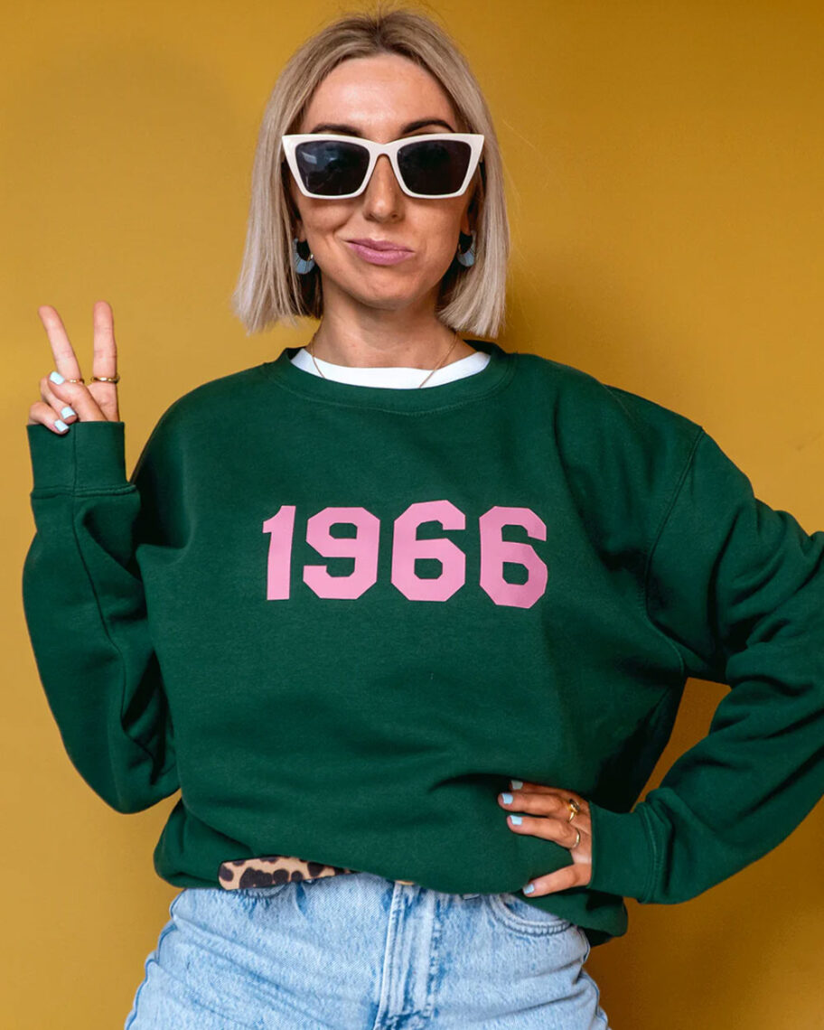 martha brook personalised stationery blog 10 unique birth year personalised birthday gifts rock on ruby personalised sweatshirt. A woman with short blonde hair poses wearing a dark green sweatshirt. The sweatshirt has '1966' written on the front in bright pink.