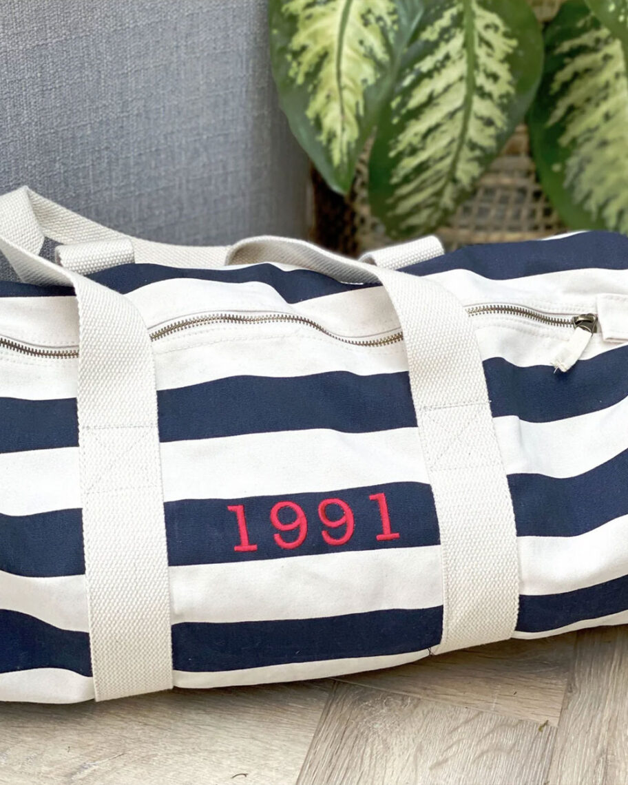 martha brook personalised stationery blog 10 unique birth year personalised birthday gifts solesmith embroidered duffle bag. A white and blue striped duffel bag on the floow next to a plant. The bag is embroidered in red with '1991'.
