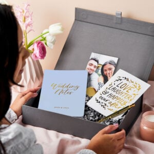 martha brook personalised luxury engagement gift box on a table covered in pink linen. The box is opened to reveal a wedding notes notebook, a gold photo holder and an engagement card that reads love laughter and happily ever after