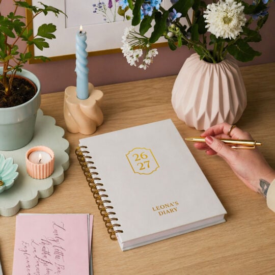 martha brook personalised stationery personalised refined 2026 2027 mid year diary hardback on a desk with a pastel blue spiral candle and spring flower arrangements