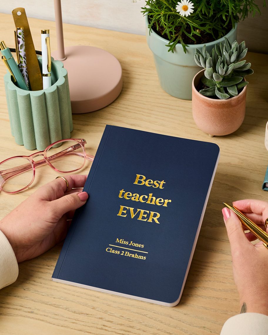 martha brook blog post thank you teacher gift guide best teacher gift softback notebook on a desk next to a pen pot, glasses being held by a pair of hands
